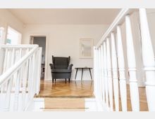 Sitzbereich in einem hellen Wohnraum mit weiß gestrichenen Wänden und Holzfußboden im Fischgrätmuster. Im Fokus steht ein grauer, gepolsterter Sessel vor einem kleinen Beistelltisch; die Szene wird von weißen Holzgeländern eingerahmt.