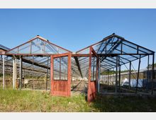 Ein verlassenes Gewächshaus mit rostigen roten Türen, die offenstehen, und einem Glasdach, das teilweise beschädigt ist. Im Hintergrund sind weitere Gewächshäuser und ein bewachsener Außenbereich zu sehen.