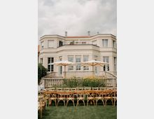 Große, helle Villa mit symmetrischer Fassade, hohen Fenstern und einer erhöhten Terrasse mit Balustrade und seitlichen Treppen. Im Vordergrund stehen auf einer Rasenfläche lange Tische mit Stühlen sowie zwei große Sonnenschirme vor einem Blumenbeet.