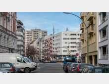 Straßenszene in einer Stadt mit mehrstöckigen Wohngebäuden unterschiedlicher Baustile, darunter ein historisches Gebäude mit roter und weißer Fassade sowie moderne Plattenbauten. Geparkte Autos säumen die Straße, und eine Ampel sowie Verkehrsschilder sind sichtbar.