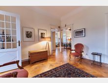 Wohnzimmer in einem Altbau mit Holzboden und hohen Decken. Der Raum ist mit einem roten Polstersessel, einer Holztruhe, einem Teppich und Wandkunst eingerichtet; im Hintergrund ein Blick in ein angrenzendes Esszimmer mit dunklen Holzstühlen.