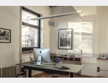 Helles, modernes Büro mit weiß gestrichenen Wänden, einem großen Fenster, das Tageslicht hereinlässt, und einem minimalistischen Schreibtisch mit einem Computer. An den Wänden hängen gerahmte Schwarz-Weiß-Fotografien, und im Hintergrund befinden sich Regale mit Dokumenten.