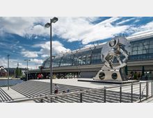 Eine moderne Glasfassade eines Bahnhofsgebäudes, flankiert von einer großen silbernen Metallskulptur, die eine abstrakte, mechanische Figur darstellt. Im Vordergrund breite Treppenstufen, Geländer und einige Personen; im Hintergrund ein Fluss und weitere städtische Gebäude.