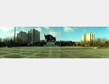 Weitläufiger Platz mit gepflastertem Boden, einer großen Statue in der Mitte, die einen Kopf mit wehender Fahne zeigt, umgeben von Bäumen und hohen Wohngebäuden im Hintergrund unter klarem Himmel.