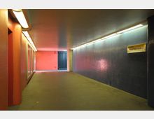 Unterführung mit farbigen Wänden, links rot und rechts mit blauen Fliesen verkleidet. Deckenbeleuchtung in Form von langen Lichtleisten; ein Schild mit der Aufschrift „Ausgang Richard-Wagner-Straße“ ist an der blauen Wand befestigt.