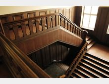 Holzverkleidetes Treppenhaus mit dekorativen Holzgeländern, breiten Stufen und großen Fenstern, die Licht in den Raum lassen. Die Architektur zeigt klassische Holzelemente und symmetrische Wandverzierungen.