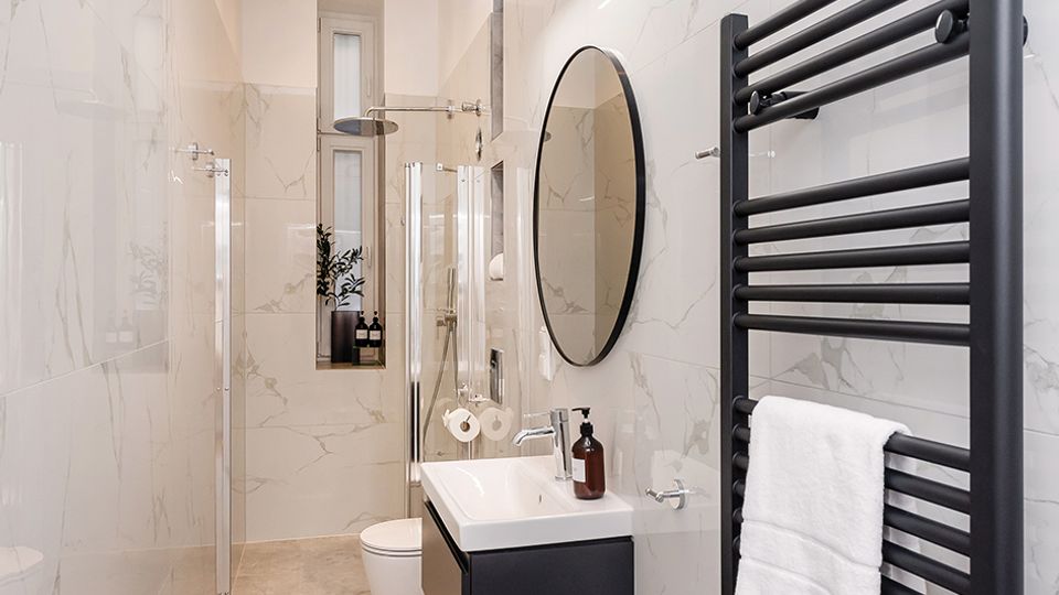 Schmales, modernes Badezimmer mit großformatigen marmorierten Wandfliesen und grauen Bodenfliesen, ausgestattet mit einer Glasdusche, wandhängendem WC und Waschtisch mit rundem Spiegel. Rechts ist ein schwarzer Handtuchheizkörper, die Decke hat eingelassene Spotlights und am Ende befindet sich ein schmales Fenster.