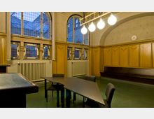 Innenraum eines historisch gestalteten Raumes mit hohen, vergitterten Fenstern und dekorativen gelben Holzpaneelen an den Wänden. In der Mitte steht ein dreieckiger Tisch mit schwarzen Stühlen, und an der Decke hängt eine Lampe mit mehreren runden, leuchtenden Glühbirnen.