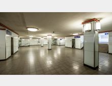 Unterirdische U-Bahn-Passage mit grauen Fliesenböden, weißen Wandfliesen und beleuchteten Säulen. Im Hintergrund befinden sich Wegweisschilder, Treppenaufgänge und eine Glaswand mit dahinterliegenden dekorativen Elementen.