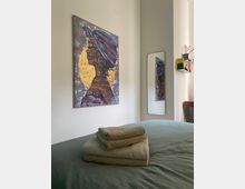 Ein Schlafzimmer mit einem gemachten Bett, auf dem gefaltete Handtücher liegen. An der Wand hängt ein farbenfrohes Gemälde einer Frau mit Kopftuch vor einem goldenen Hintergrund; daneben steht ein schmaler Spiegel, der das Bild reflektiert.