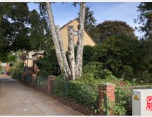 Straßenszene mit einem Wohnhaus im Hintergrund, umgeben von dichtem Grün. Im Vordergrund befindet sich ein Gehweg, ein Zaun aus Metall und roten Backsteinsäulen sowie ein Stromkasten mit Graffiti.