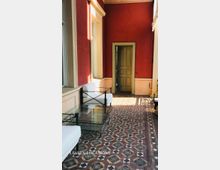 Überdachter Korridor mit roten Wänden und hellen Stuckrahmen, einem alten Holzportal am Ende und einem dekorativen Fliesenboden mit geometrischem Muster. Entlang der Fensterfront stehen weiß gepolsterte Sitzmöbel und ein gläserner Couchtisch mit Metallgestell.