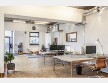 Modernes Büro mit offenem Grundriss, großen Fenstern für Tageslicht, minimalistischen Möbeln und sichtbaren Lüftungsrohren an der Decke. Im Raum befinden sich mehrere Arbeitstische, ein Besprechungsbereich mit Teppich und Sitzmöbeln sowie Pflanzen als Dekoration.