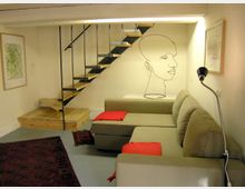 Wohnbereich in einem modernen Stil mit einer schmalen, offenen Holztreppe, die an Metallstreben befestigt ist. Neben einem beigefarbenen Sofa mit roten Kissen befinden sich minimalistische Wanddekorationen, darunter eine Konturzeichnung eines Gesichts, sowie ein Stehlampe und ein orientalischer Teppich auf dem Boden.