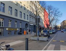 Straßenszene in einer Stadt mit mehrstöckigen Wohngebäuden, teilweise in grauer und roter Farbe gestrichen. Im Erdgeschoss befinden sich Geschäfte, darunter ein Nagelstudio mit der Aufschrift „HELLO NAILS & SPA“. Parkende Autos und kahle Bäume säumen die Straße.