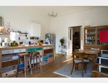 Wohnküche mit Holzfußboden, einer Küchenzeile aus gemusterten Schubladen und offenen Regalfächern, zwei Barhockern, einem Esstisch mit Holzstühlen und einem hohen Holzschrank mit Glastüren. Im Hintergrund befinden sich eine Tür mit Blick in einen Nebenraum sowie ein Kühlschrank.