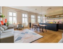 Helles, modern eingerichtetes Wohnzimmer mit offener Küche. Der Raum verfügt über große, bogenförmige Fenster, eine graue Couch, einen farbigen Teppich, einen Esstisch aus Holz mit Stühlen und eine schwarze Küchenzeile mit Regalen, die Gläser und Flaschen enthalten.