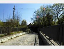 Pflasterstraße mit Geländern auf beiden Seiten, die zu einer kleinen Unterführung unter einer Brücke führt. Die Umgebung ist von Bäumen und Sträuchern geprägt, im Hintergrund sind Bahngleise sowie ein Strommast sichtbar.