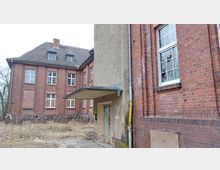 Backstein-Gebäude im verfallenen Zustand, mit zahlreichen Fensteröffnungen und teils zerschlagenen Scheiben; ein Anbau mit Putzfassade und einem kleinen Vordach ragt hervor, umgeben von verwildertem Gelände.