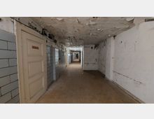 Langer, verlassener Korridor mit abblätternder Farbe an den Wänden und der Decke sowie freiliegenden Kabeln. Die Wände sind teilweise mit hellen Fliesen verkleidet, und eine schlichte Holztür befindet sich links im Vordergrund.