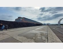 Industriegelände mit einer großen Lagerhalle aus Metall und Glas, auf der "Mebra Remine" steht. Rechts verlaufen Bahngleise und es sind Kräne im Hintergrund sichtbar, während links hohe Kohle- oder Schutthaufen zu sehen sind.