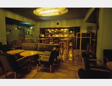 Barbereich eines gemütlichen Lokals mit Holzboden, dunklen Polstermöbeln und runden Holztischen. Im Hintergrund ist eine beleuchtete Theke mit Flaschenregalen zu sehen, über der mehrere Hängelampen angebracht sind.