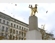 Eine goldene Skulptur von zwei tanzenden Figuren steht auf einem hohen Betonpfeiler, umgeben von gelb belaubten Bäumen. Im Hintergrund ist die Fassade eines historischen Gebäudes mit vielen Fenstern zu sehen, während einige Vögel am wolkigen Himmel fliegen.