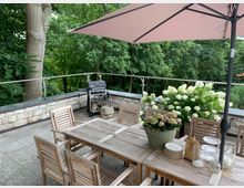 Terrasse mit Holzmöbeln, einem großen Esstisch und Stühlen, darüber ein rosa Sonnenschirm. Im Hintergrund befinden sich eine Steinbrüstung mit Metallgeländer, eine Gruppe von Bäumen sowie weiße Hortensien in Pflanzkübeln. Ein Grill steht an der Brüstung.