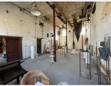 Innenansicht eines Kunstateliers in einem ehemaligen Industriegebäude mit weiß gestrichenen Ziegelwänden, freiliegender Holzbalkendecke und großen Fenstern. Der Raum ist mit Skulpturen, Sockeln und Kunstwerken eingerichtet, die verteilt auf Metallgestellen und Tischen präsentiert werden.