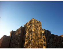 Wohnkomplex aus Beton mit mehreren hohen, rechteckigen Gebäuden, deren Fassaden zahlreiche Fenster enthalten, im Sonnenlicht beleuchtet; dahinter ein klarer blauer Himmel.