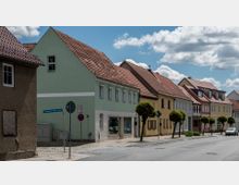 Straße mit Reihen von Wohnhäusern im traditionellen Stil, teils verputzt und in Pastellfarben gestrichen, mit roten Ziegeldächern. Vor den Häusern stehen kleine Bäume, und das Straßenschild weist auf die "Geschwister-Scholl-Straße" hin.