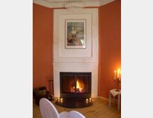 Ein gemütlicher Raum mit einem weißen Kamin, in dem ein Feuer brennt. Über dem Kamin hängt ein gerahmtes Poster, die Wände sind in Orange gehalten, und ein Kerzenständer steht auf einem kleinen Tisch daneben.