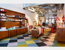 Ein Ladeninnenraum mit buntem, geometrisch gemustertem Boden und einer modernen Einrichtung. Holzregale und Kommoden an den Wänden sind mit verschiedenen Produkten wie Textilien, Kosmetika und Dekorationsartikeln bestückt, während die Decke mit sichtbaren Lüftungsrohren gestaltet ist.