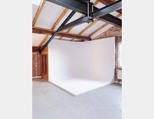 Ein Fotostudio mit gebogener, weißer Hohlkehle, freiliegenden Ziegelwänden, hölzernen Deckenbalken und schwarzen Metallträgern an einer hohen Decke. Der Raum hat einen Betonboden und ein Fenster auf der rechten Seite.