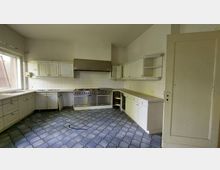 Helle Küche mit weißen Schränken, Edelstahlherd samt großer Dunstabzugshaube und gefliestem Spritzschutz. Der Boden besteht aus blauen Fliesen in einem geschwungenen Muster, neben der Tür und einem Fenster mit Blick nach draußen.