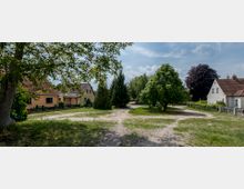 Dorfplatz mit vereinzelten Wohnhäusern mit roten Ziegeldächern, unbefestigten Wegen und Bäumen sowie Sträuchern, die den Platz umgeben. Im Vordergrund führt ein sandiger Weg durch eine grüne Wiese.