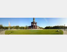 Panorama-Aufnahme der Siegessäule in Berlin, umgeben von einer Grünfläche und Bäumen. Der Himmel ist klar, und im Vordergrund sind Gehwege und eine Fahne sichtbar.