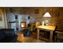 Gemütliches Wohnzimmer mit gemauerter Kaminecke, die ein brennendes Feuer zeigt, und einer Sitzgruppe mit Holztisch und Polsterstühlen. Die Wände aus sichtbarem Backstein, hölzernes Parkett und eine Hängelampe tragen zur warmen Atmosphäre bei.