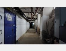 Ein langer Kellerflur mit weißen, teils fleckigen Wänden, sichtbar freiliegenden Rohren an der Decke und vereinzelten Lichtquellen. Auf der linken Seite befindet sich eine blaue Metalltür mit der Aufschrift "TAV2 STOER RESERVE", rechts sind verschiedene Gegenstände und Maschinen gelagert.