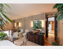 Ein modern eingerichtetes Badezimmer mit Bambus-verkleideten Wänden, dunklem Holzwaschtisch mit Aufsatzwaschbecken, freistehender Badewanne und einem großen Spiegel. Grünpflanzen und Holzdetails sorgen für eine natürliche Atmosphäre, während eine Tür zu einem angrenzenden Raum mit Holzboden führt.