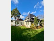 Einfamilienhaus mit dunkel gedecktem Walmdach und weißer Fassade, angrenzend an einen flachen Anbau mit großer Glasfront und Terrasse. Im Vordergrund liegt ein gepflegter Rasen mit Sträuchern und hohen Bäumen unter blauem Himmel.