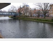 Ein Fluss mit steinernen Ufern verläuft durch eine Stadtlandschaft, flankiert von kahlen Bäumen und Backsteingebäuden. Eine Fußgängerbrücke aus Stahl und Glas überspannt den Fluss, während im Hintergrund mehrstöckige Häuser und Wandkunst zu sehen sind.