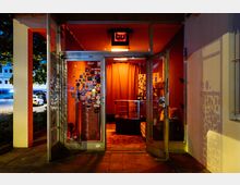 Eingang eines kleinen, rot-orange beleuchteten Geschäfts oder Veranstaltungsraums mit verglasten Türen, die mit Aufklebern bedeckt sind. Der Innenraum zeigt Vorhänge, eine Vitrine und Poster im Hintergrund, während die Umgebung eine städtische Straßenszene mit geparkten Autos erkennen lässt.