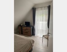 Ein kleines Schlafzimmer mit schräger Decke, einem Bett mit heller Bettdecke, einer Kommode in Holzoptik mit einem kleinen Fernseher darauf, sowie einer Glastür mit halbtransparenten Vorhängen, die auf einen Balkon führt.