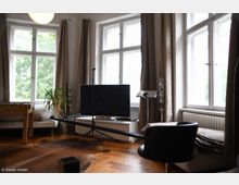 Wohnzimmer mit großen weißen Fenstern, beigefarbenen Vorhängen und einem Holzboden. Im Raum stehen ein Flachbildfernseher, Lautsprecher, ein Sofa, ein schwarzer Stuhl, ein Kickertisch und eine Zimmerpflanze, darunter liegt ein Teppich mit Fell-Optik.