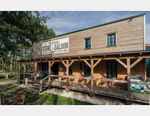 Holzgebäude im Westernstil mit der Aufschrift „The Buchholz Store & Saloon“ auf einem Schild an der Fassade. Das Gebäude hat eine überdachte Veranda mit Holzstützen, auf der rote Stühle und ein kleines Ben & Jerry's Werbeschild stehen, umgeben von Bäumen und einer Wiese.