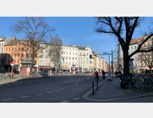 Straßenszene in einem urbanen Viertel mit mehrstöckigen Wohngebäuden in verschiedenen Farben, darunter ein orangefarbenes und ein pastellfarbenes Gebäude. Im Vordergrund sind ein breiter Gehweg mit geparkten Fahrrädern, ein Fahrradweg und eine Kreuzung mit Fußgängern und Radfahrern zu sehen, während kahle Bäume die Umgebung säumen.