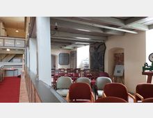 Innenraum einer historischen Kirche oder Kapelle mit bemalten Holzbalken, einem roten Teppichboden und einer Sitzgruppe aus unterschiedlich farbigen Stühlen. An den Wänden hängen Tafeln und Informationen, darunter eine große beschriftete Tafel mit Vogel-Motiv, sowie ein Tisch mit Prospekten.