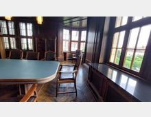 Raum im historischen Stil mit Holztäfelungen, einem großen Tisch und Stühlen mit Lederpolsterung. Große Fenster mit Sprossenrahmen bieten Blick auf rote Dächer und Grünflächen außerhalb des Gebäudes.