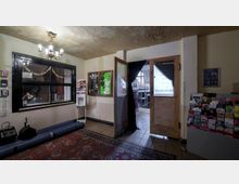 Ein Eingangsbereich eines Veranstaltungsraums mit einer gläsernen Durchreiche, einem Kronleuchter an der Decke, mehreren Plakaten an der Wand sowie einer Prospektablage auf der rechten Seite. Eine Tür mit der Aufschrift „Ballhaus Ost“ führt zu einem Raum mit Stuhlreihen und einer großen Fensterfront.